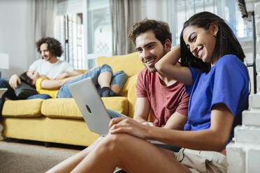 Smiling young couple sharing laptop while friends relaxing on sofa in living room at home - JSMF01596