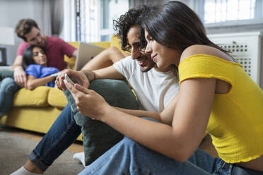 Young couple sharing digital table while friends relaxing in living room at home - JSMF01597