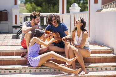 Fröhliche junge Freunde, die mit einer Gitarre auf den Stufen einer Terrasse sitzen und sich an einem sonnigen Tag vergnügen - JSMF01632