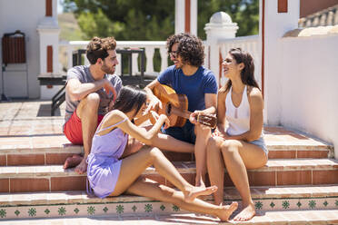 Fröhliche junge multiethnische Freunde, die mit einer Gitarre auf den Stufen einer Terrasse sitzen und sich an einem sonnigen Tag vergnügen - JSMF01633