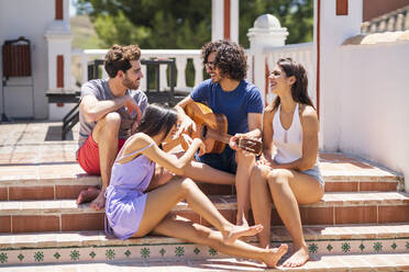 Happy young multi-ethnic friends enjoying while sitting with guitar on steps at terrace during sunny day - JSMF01633