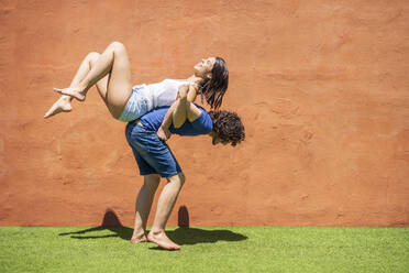 Young man carrying woman on back by brown wall during sunny day - JSMF01634