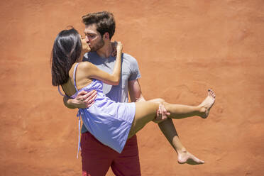 Young man kissing while carrying woman against brown wall at back yard during sunny day - JSMF01638