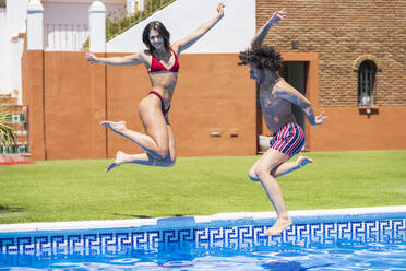 Glückliches junges Paar, das an einem sonnigen Tag in den Swimmingpool im Hinterhof springt - JSMF01645