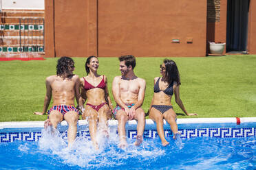 Fröhliche junge multiethnische Freunde spritzen mit Wasser, während sie an einem sonnigen Tag am Pool sitzen - JSMF01647