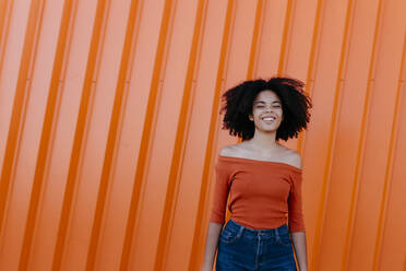 Fröhliche junge Afro-Frau, die an einer orangefarbenen Metallwand steht - TCEF00913