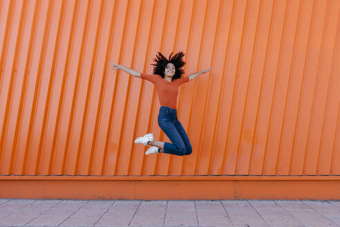 Happy young woman jumping with arms outstretched against orange wall - TCEF00914