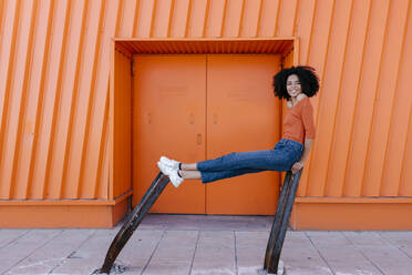 Smiling young afro woman sitting on damaged metal against orange door - TCEF00915