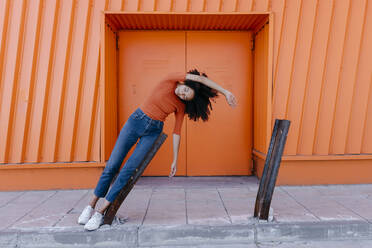 Junge Frau lehnt an beschädigtem Metall mit erhobenem Arm gegen eine orangefarbene Tür - TCEF00918