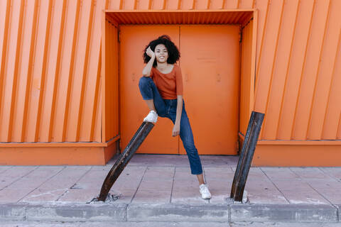 Smiling young afro woman leaning on metal with hand in hair against orange door stock photo