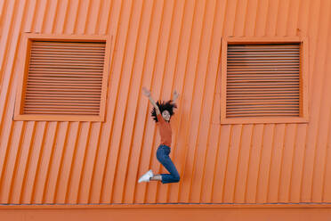 Happy young woman jumping with arms raised against orange wall - TCEF00926