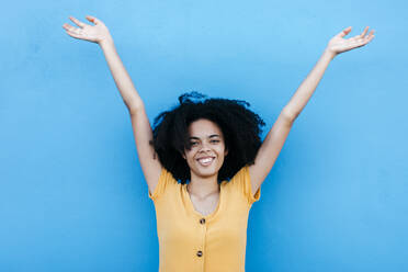 Glückliche junge Afro-Frau, die mit erhobenen Armen an einer blauen Wand steht - TCEF00929
