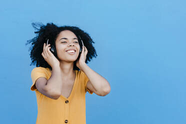 Young afro woman enjoying while listening music through headphones against blue wall - TCEF00932