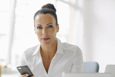 Confident businesswoman using smart phone while sitting in home office - JOSEF01276