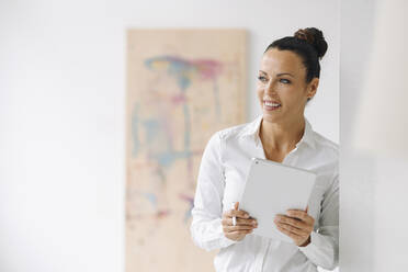 Smiling businesswoman holding digital tablet looking away while standing by wall in home office - JOSEF01282