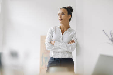 Thoughtful businesswoman with arms crossed standing by wall in home office - JOSEF01283