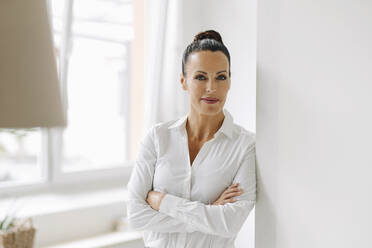 Confident female entrepreneur with arms crossed standing by wall in home office - JOSEF01287