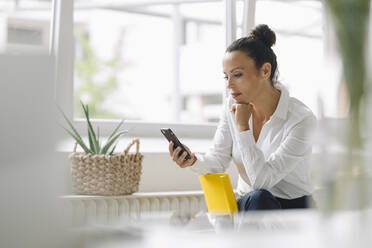 Eine Unternehmerin benutzt ein Mobiltelefon, während sie in ihrem Büro am Fenster sitzt - JOSEF01293
