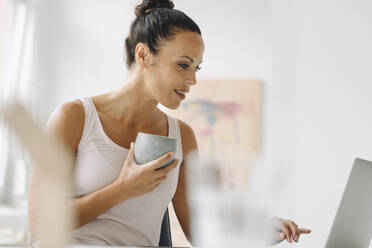 Woman holding coffee cup using laptop while sitting against wall at home office - JOSEF01324