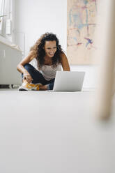 Fröhliche Frau im mittleren Erwachsenenalter, die einen Laptop benutzt, während sie zu Hause auf dem Boden sitzt - JOSEF01332