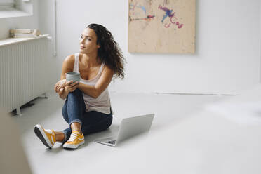 Thoughtful woman holding coffee mug while sitting with laptop on floor at home - JOSEF01340