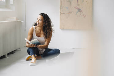 Smiling mid adult woman holding book looking away while sitting on floor at home - JOSEF01346