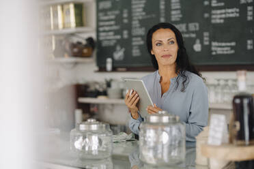 Smiling female owner holding digital tablet contemplating while standing in cafe - JOSEF01403