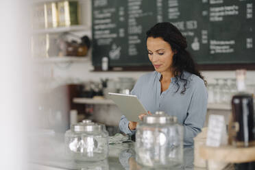 Mid adult woman using digital tablet while standing at counter in coffee shop - JOSEF01404