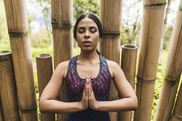 Junge Frau meditiert mit geschlossenen Augen und an Bambus geklammerten Händen im Park - DSIF00016