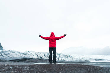 Rückenansicht einer Person in warmer Kleidung, die mit ausgestreckten Armen in einer malerischen Schneelandschaft in Skaftafell, Island und Vatnajokull steht - ADSF00422