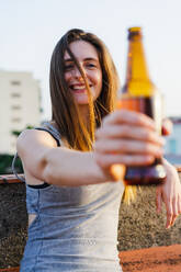 Schöne, lebhafte, lächelnde junge Frau, die eine Bierflasche in der ausgestreckten Hand hält und in die Kamera schaut, während sie sich bequem auf eine Brüstung mit einer verschwommenen Stadt im Hintergrund stützt - ADSF00424