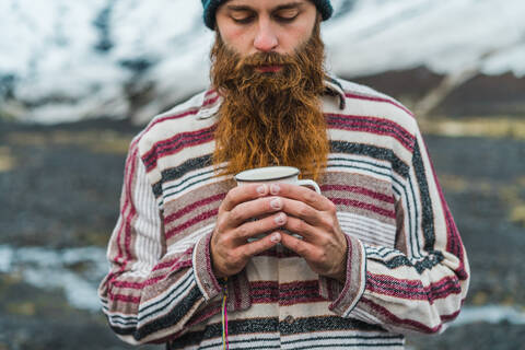 Attraktiver bärtiger Mann, der einen Becher mit einem heißen Getränk in der Hand hält und wegschaut, während er sich an einen Metallzaun lehnt, nicht weit von einer kleinen Hütte in der isländischen Landschaft, lizenzfreies Stockfoto
