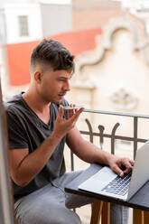 Adult handsome man sitting on balcony watching laptop and sending voice message via smartphone - ADSF00450