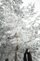 Hübsche junge Frau in stilvollem Outfit, die wegschaut, während sie in der Nähe eines mit Schnee bedeckten Baumes an einem kalten Tag in einer wunderschönen Landschaft steht - ADSF00492