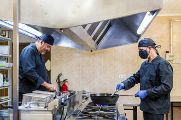 Traditional cooking in restaurant kitchen, chef wearing protective mask - DLTSF00843