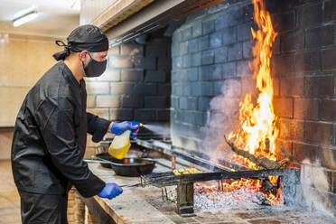 Traditional cooking of paella in restaurant kitchen, chef wearing protective mask - DLTSF00846