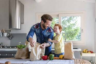 Cheerful father and son with groceries standing at dining table in kitchen - EIF00015