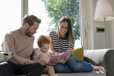 Lächelnde Frau, die ein Bilderbuch liest, während sie neben ihrem Sohn und einem Mann auf dem Sofa im heimischen Wohnzimmer sitzt - EIF00040