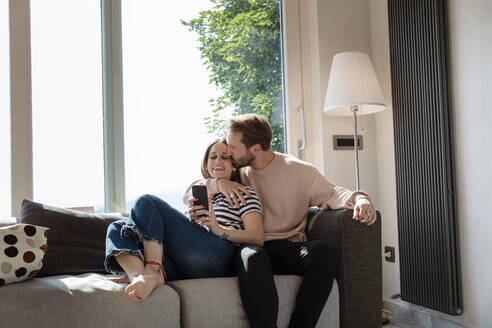 Mann küsst Frau, während er sich auf dem Sofa vor dem Fenster im Wohnzimmer ausruht - EIF00046