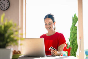 Lächelnde schöne Frau, die eine Kreditkarte hält, während sie einen Laptop am Esstisch vor dem Fenster benutzt - EIF00051