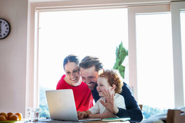 Lächelnde Familie bei einem Videogespräch über den Laptop am Esstisch vor dem Fenster - EIF00053