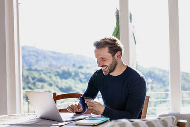 Lächelnder gut aussehender Mann, der eine Kreditkarte hält, während er einen Laptop am Esstisch vor dem Fenster benutzt - EIF00055