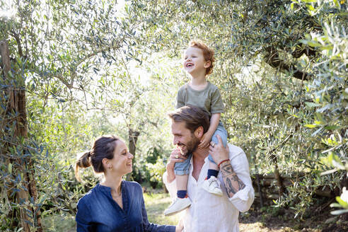 Lächelnde Eltern, die sich gegenseitig ansehen, während sie ihren Sohn auf den Schultern durch einen Olivenhain tragen - EIF00062