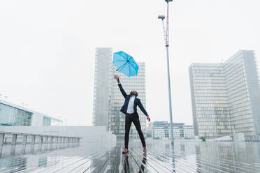 Afroamerikanischer Mann in Geschäftskleidung, der versucht, einen blauen Regenschirm aufzufangen, der von einem Windstoß hochgeschleudert wird, und der nach oben schaut, während er auf dem nassen Bürgersteig vor dem Hintergrund grauer Stadtgebäude steht - ADSF00510