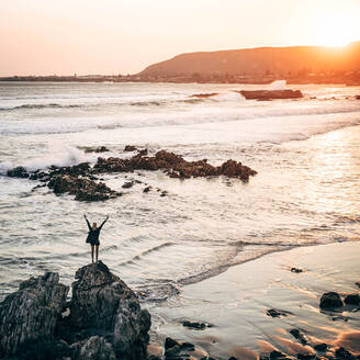 Rückenansicht einer Touristin, die mit gespreizten Händen auf einem Stein an der Küste steht und die Freiheit am Meer spürt. - ADSF00532
