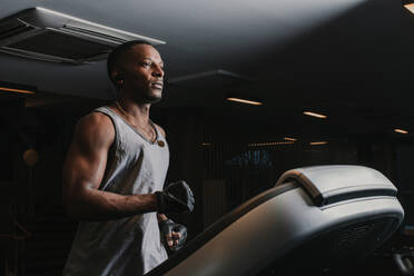 Side view of handsome African American guy in sportswear listening to music and running on treadmill while training in modern gym - ADSF00547