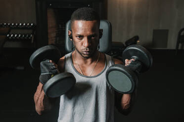 Young African American man lifting two heavy dumbbells and listening to music during training in modern gym - ADSF00560