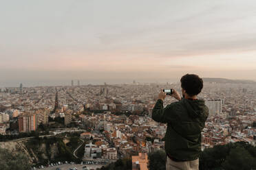 Back view of man in casual outfit using modern smartphone to take pictures of amazing metropolitan city in wonderful cloudy evening - ADSF00597