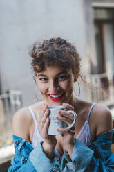 Junges lockiges Mädchen in weißer Unterwäsche und Jeansjacke steht mit Kaffeetasse auf dem Balkon und schaut verträumt in die Ferne - ADSF00605