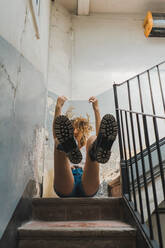 Von unten von einem lockigen Mädchen in Shorts und Stiefeln, das auf einer Treppe sitzt, mit fliegenden Locken und lachend in Bewegung - ADSF00611
